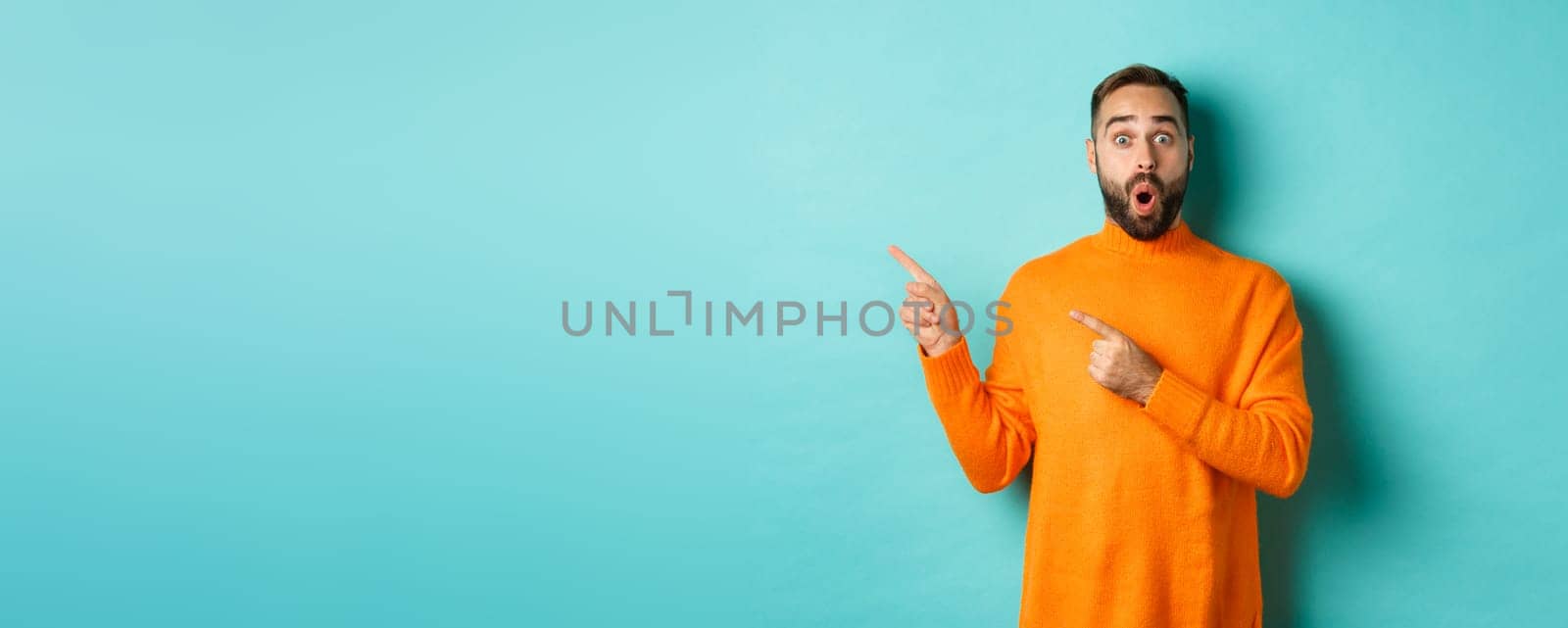 Impressed young man with beard, reacting to promo banner, pointing fingers left, showing logo and looking surprised, standing over turquoise background by Benzoix