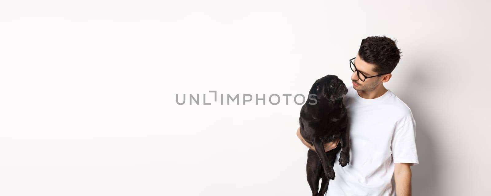 Handsome young man holding his black pug, dot and pet owner looking each other faces, standing against white background.