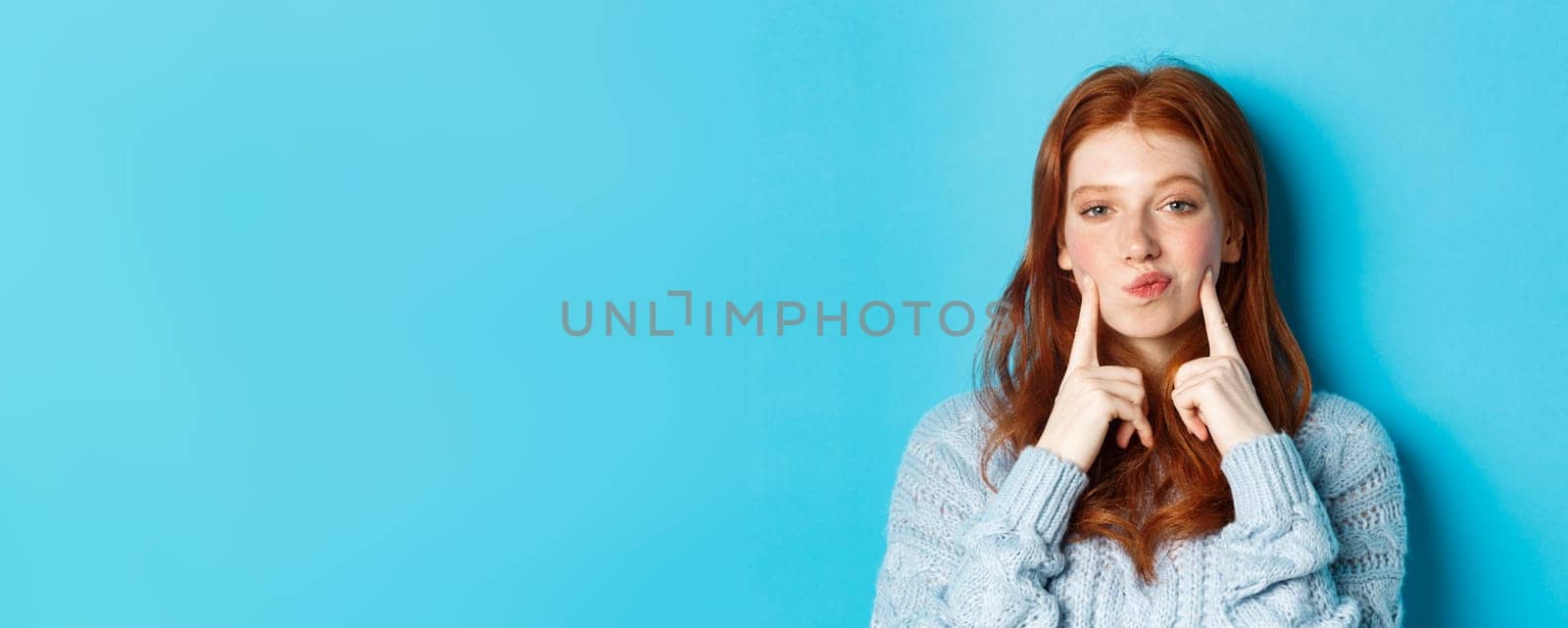 Close-up of cute redhead girl in sweater, pucker lips and pointing fingers at cheeks, poking dimples, standing over blue background by Benzoix