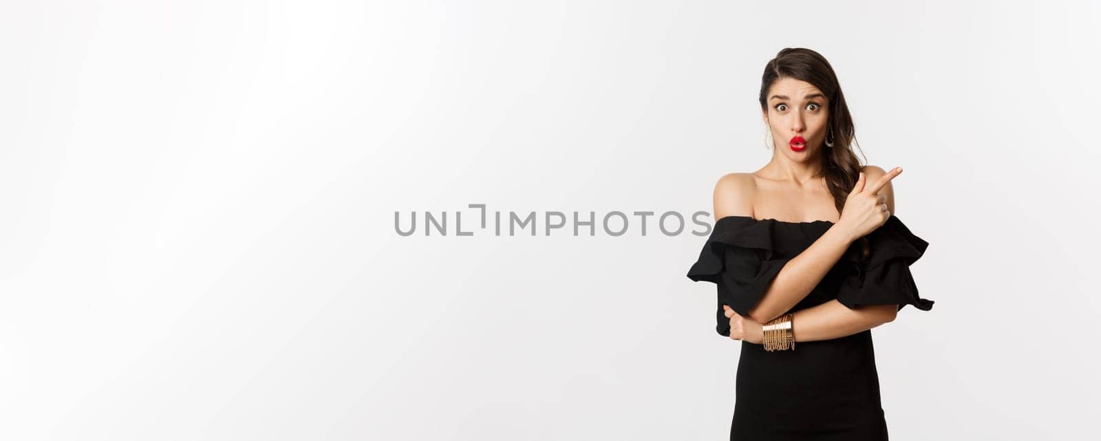 Fashion and beauty. Surprised woman in black dress pointing finger at upper right corner, showing advertisement, standing over white background by Benzoix