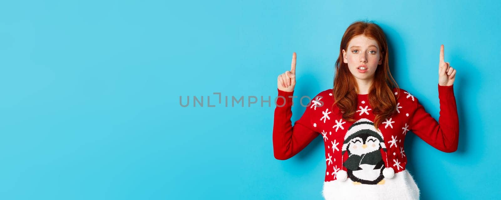 Merry Christmas. Skeptical and unamused redhead girl pointing fingers up, showing logo with reluctant face, standing over blue background by Benzoix