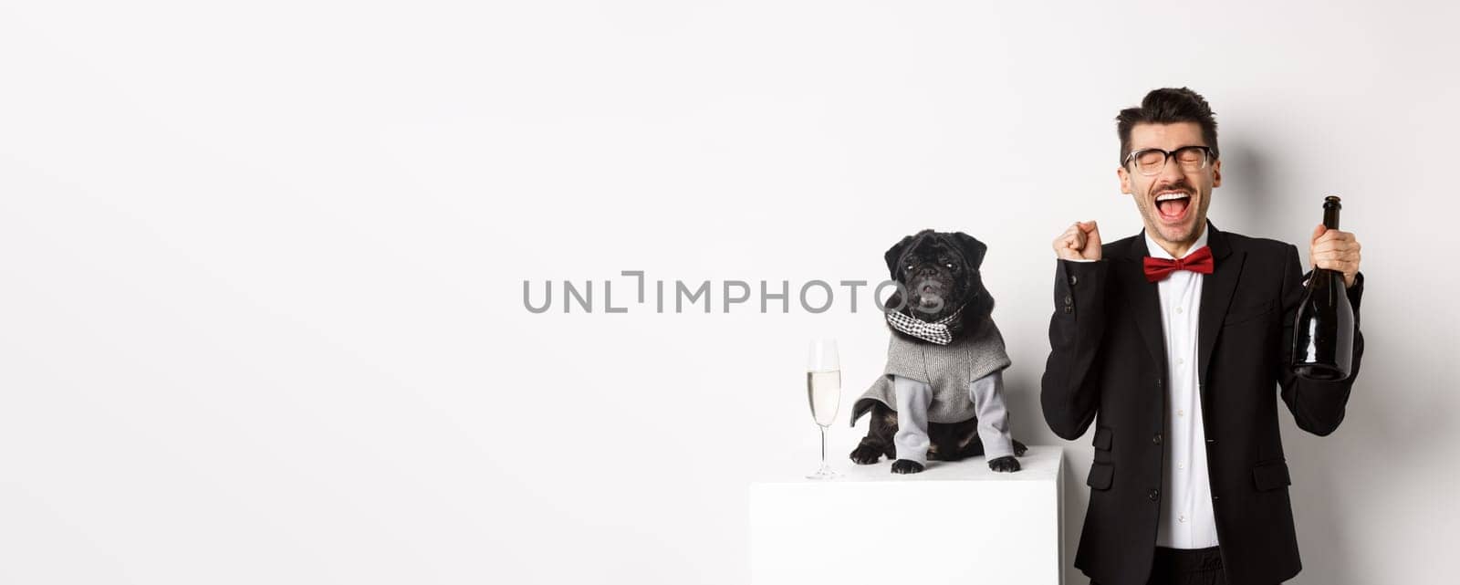 Pets, winter holidays and New Year concept. Happy young man celebrating Christmas with cute black dog wearing party costume, holding bottle champagne, white background.