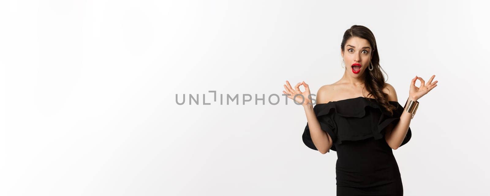 Fashion and beauty. Attractive brunette woman in black dress, showing okay signs and staring excited, approve and recommend, standing over white background by Benzoix