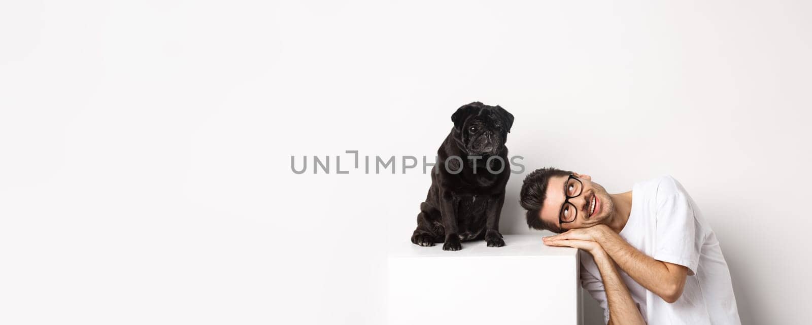 Handsome young man lay head near cute black pug, smiling and looking up at copy space, white background.