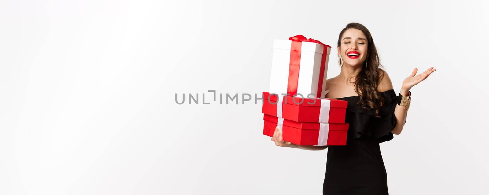 Celebration and christmas holidays concept. Excited and happy woman receive gifts, holding xmas presents and rejoicing, standing in black dress over white background.