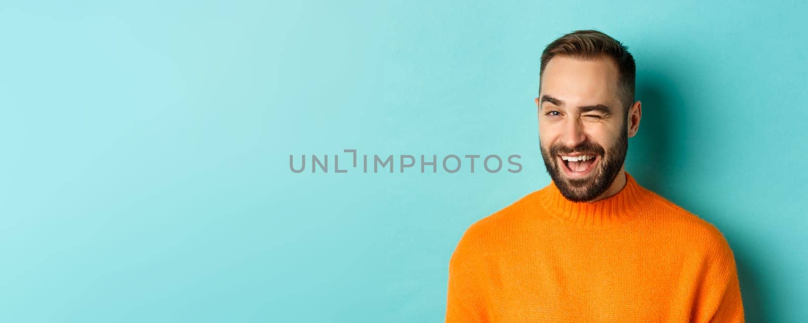 Handsome confident man winking at camera, smiling sassy, standing in orange sweater against blue background by Benzoix