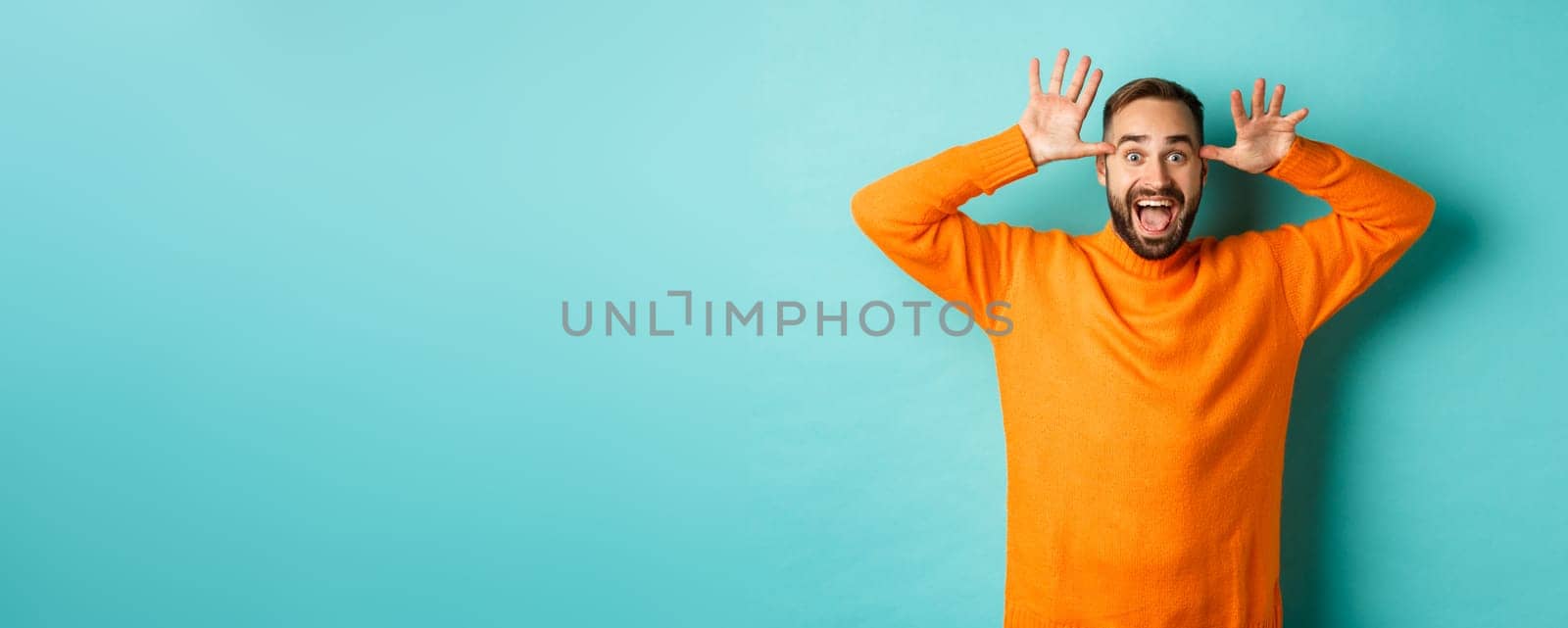 Image of handsome caucasian man making funny faces, mocking someone and smiling, standing against light blue background.
