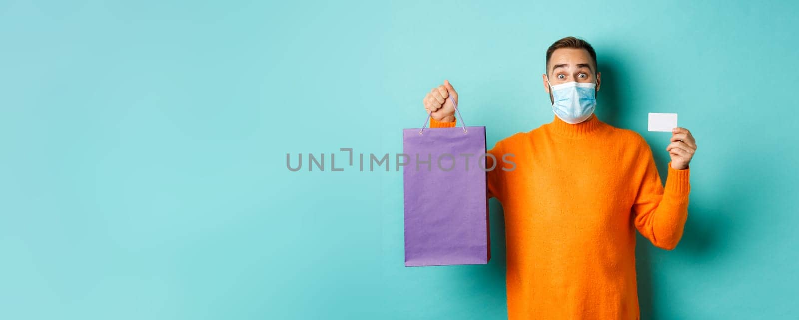 Covid-19, pandemic and lifestyle concept. Happy male customer showing credit card and purple shopping bag, standing over light blue background.