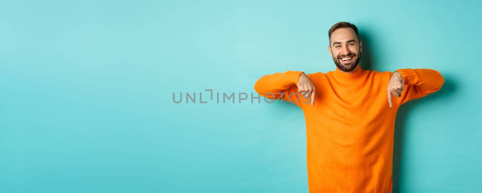Happy adult man with beard pointing fingers down, smiling cheerful, showing advertisement, standing over turquoise background by Benzoix