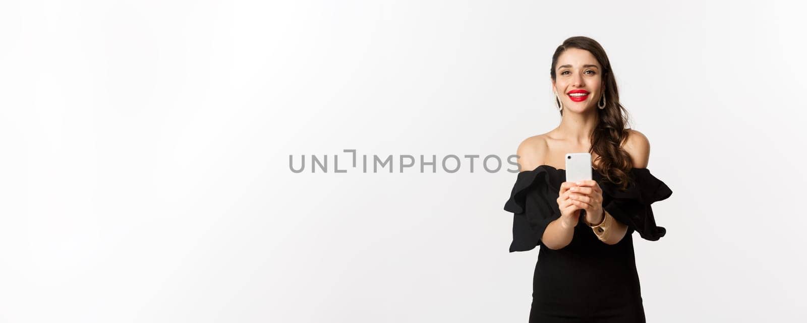 Attractive party girl taking photo on smartphone, make photograph on mobile phone camera, standing in black dress over white background by Benzoix