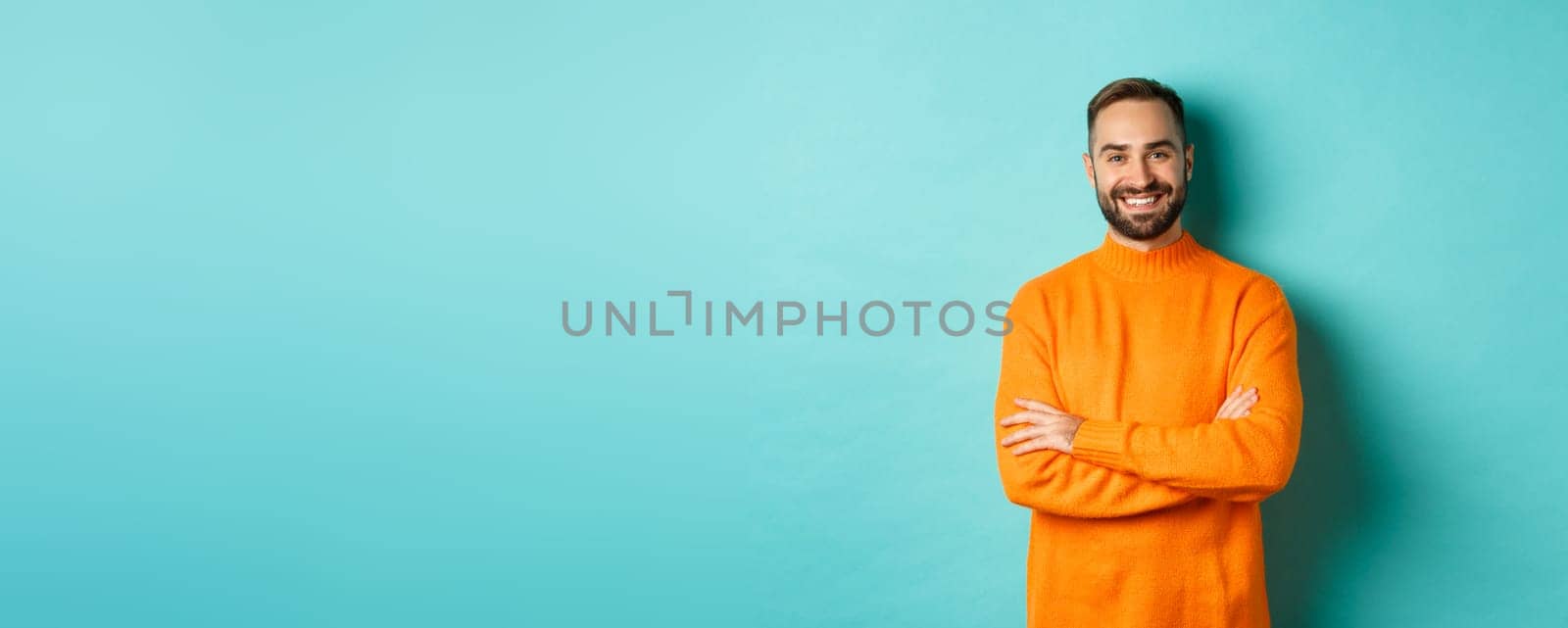 Handsome bearded male model in orange sweater, looking confident and smiling, cross arms on chest, standing over turquoise background.