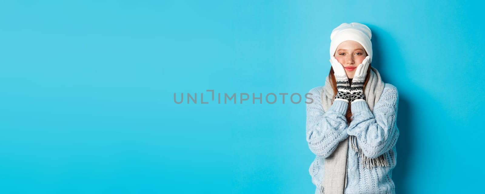 Winter and holidays concept. Cute girl in white beanie, sweater and gloves, squeezing cheeks and smiling pleased, warm-up after cold outdoors, standing over blue background.