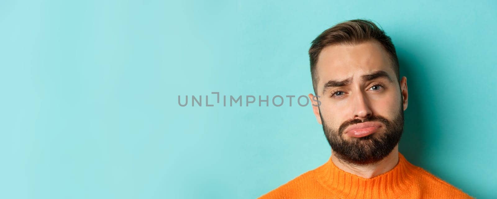 Headshot of sad and gloomy man complaining, pouting and frowning disappointed, standing against turquoise background.