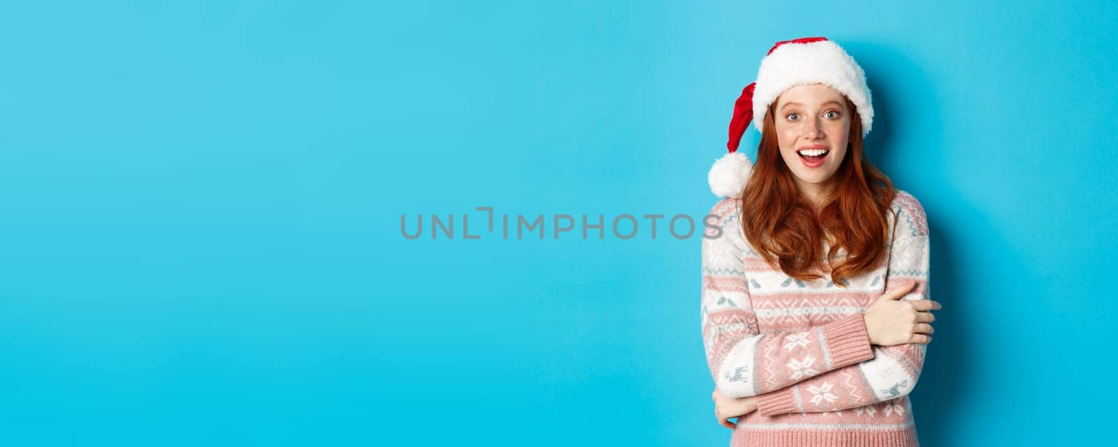 Winter and Christmas Eve concept. Amazed redhead girl in santa hat staring at camera in awe, smiling excited, standing over blue background.