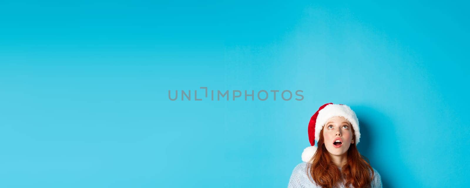 Winter holidays and Christmas eve concept. Head of pretty redhead girl in santa hat, appear from bottom and looking up at logo, seeing promo offer amazed, blue background.