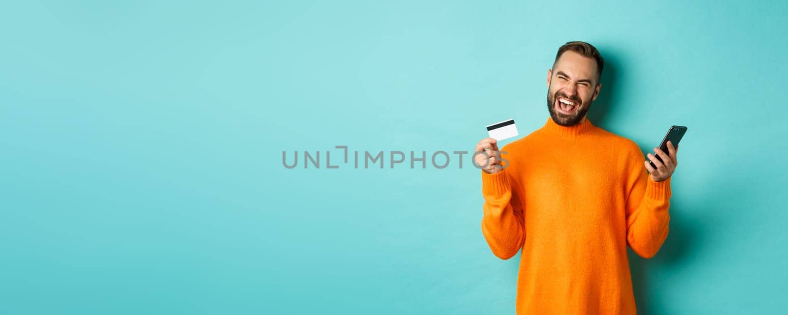 Online shopping. Happy young man using mobile phone and credit card, paying internet, light blue background.