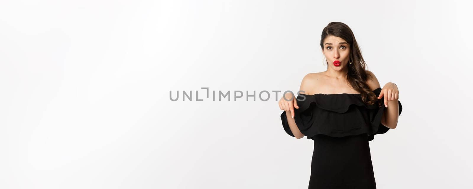 Fashion and beauty. Elegant woman in black dress pointing fingers down, showing promo and smiling, standing over white background.