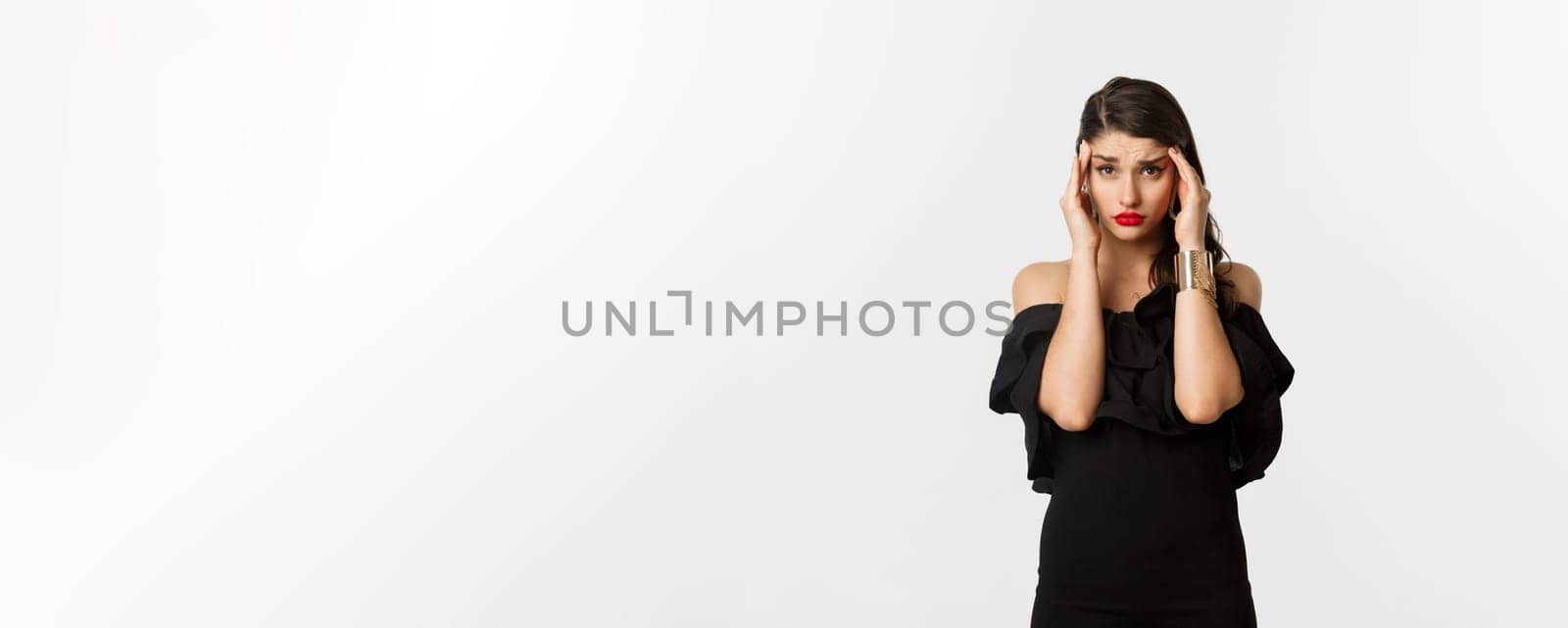 Fashion and beauty. Young modern woman in glamour dress, jewelry and makeup, touching head and looking exhausted, feeling dizzy, standing over white background.