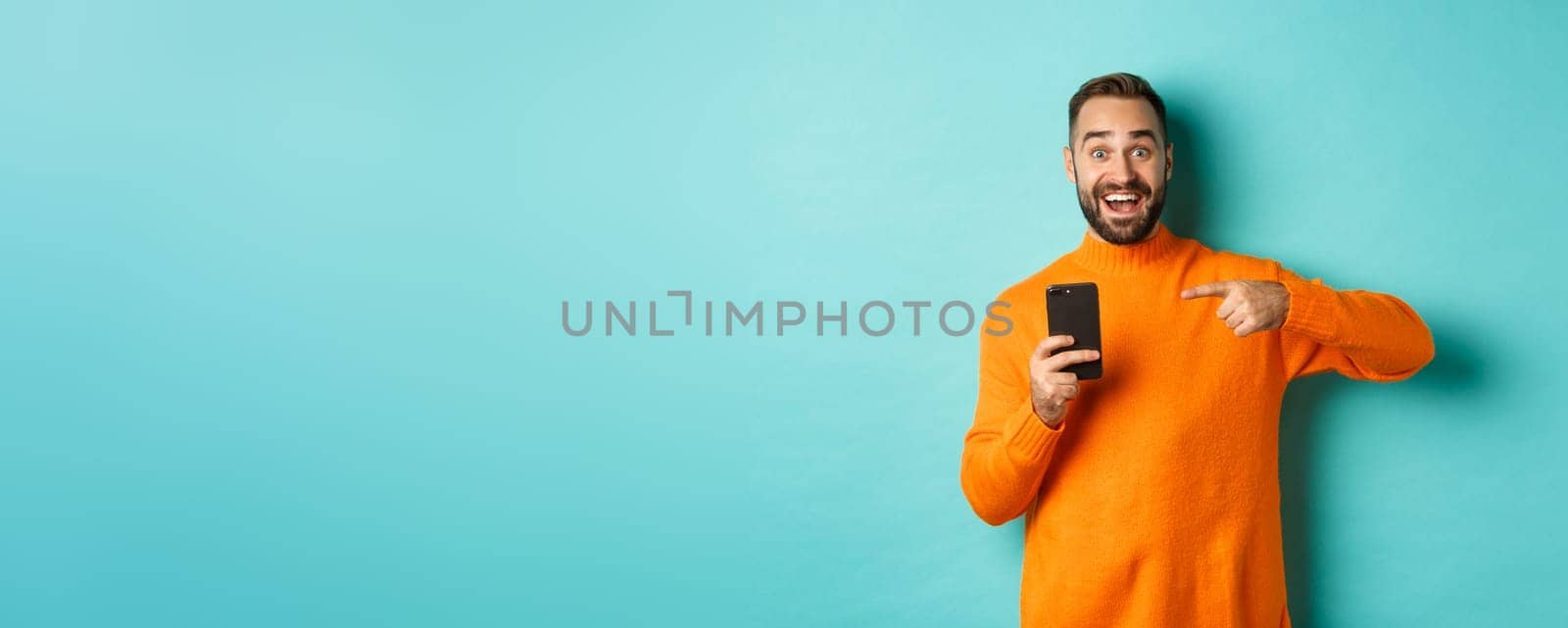 Excited man pointing at mobile phone, showing internet promo, standing over turquoise background, concept of online shopping.