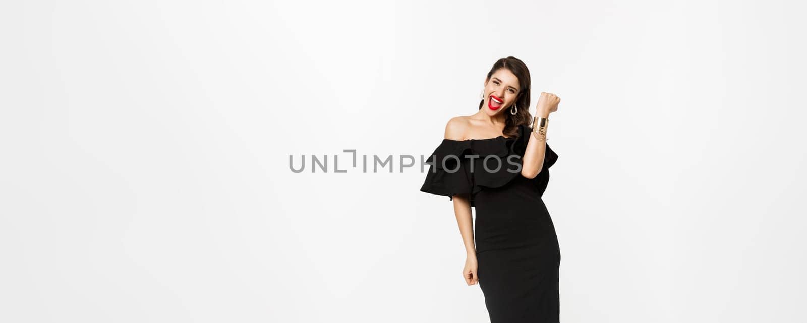 Beauty and fashion concept. Full length shot of happy young woman in luxury black dress, jewelry and heels, wearing makeup and looking satisfied, celebrating victory, winning, white background.