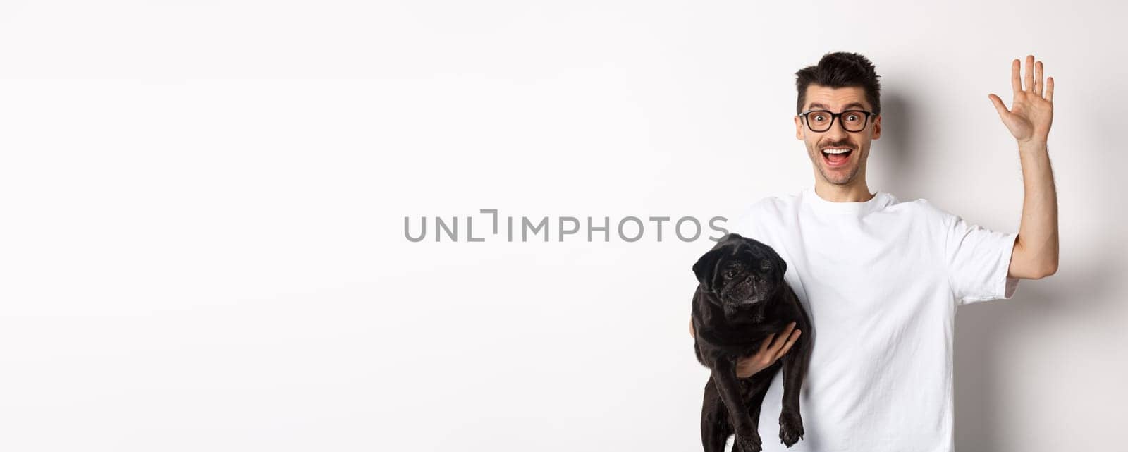 Handsome young man in glasses holding his black pug and waving hand, guy saying hello while carry dog with one arm, standing over white background.