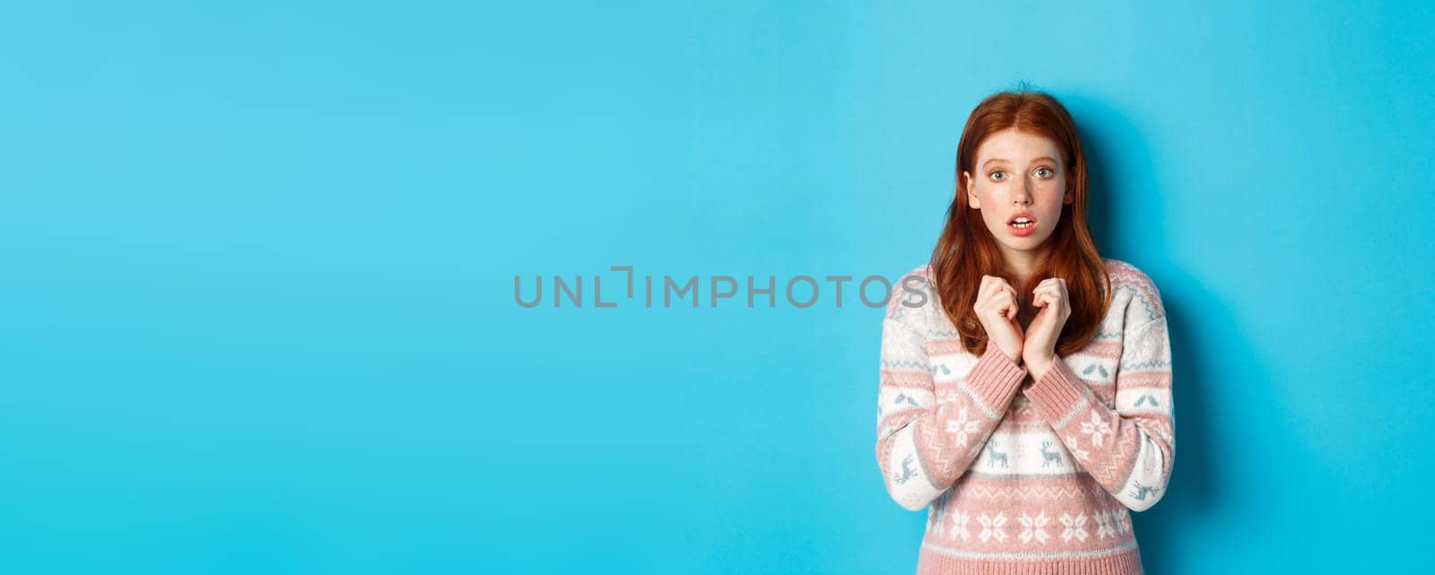 Close-up of cute redhead girl looking with anticipation and worry, staring at camera, standing in winter sweater against blue background by Benzoix