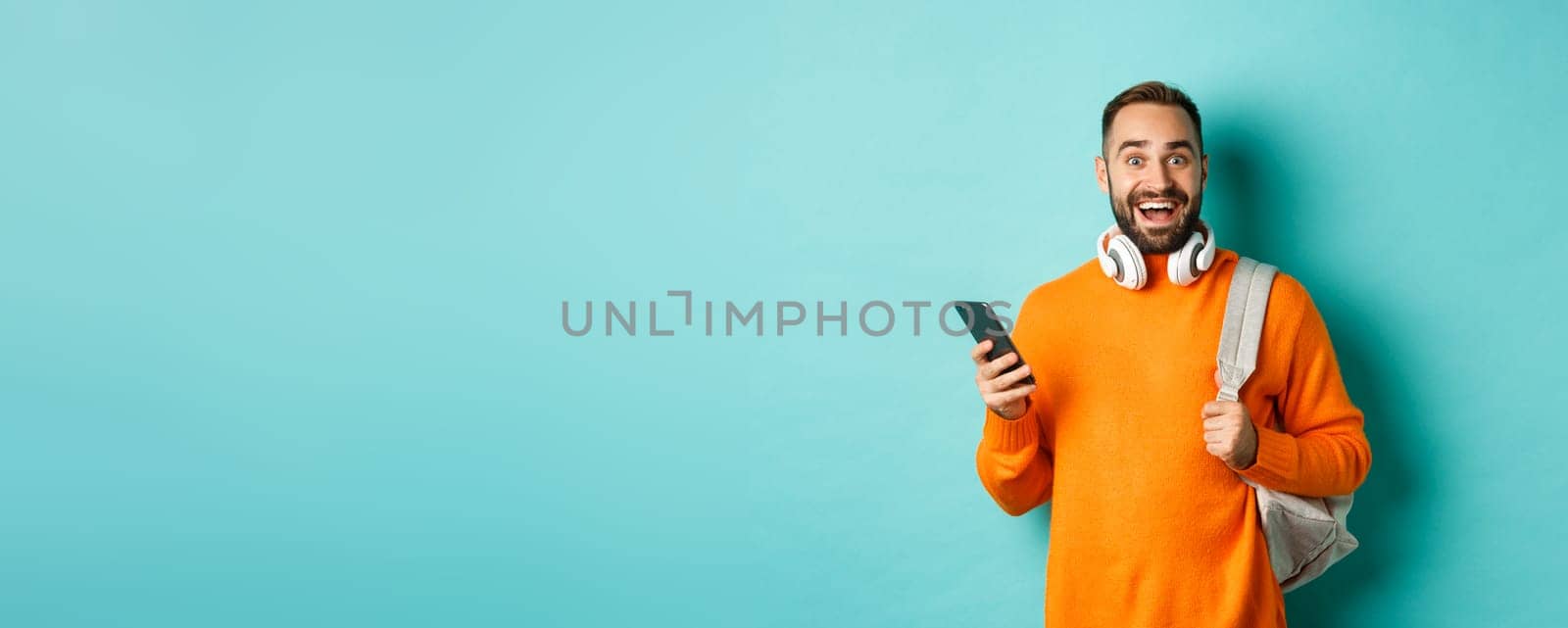 Caucasian man with headphones and backpack staring at camera amazed after reading phone notification, standing over turquoise background by Benzoix