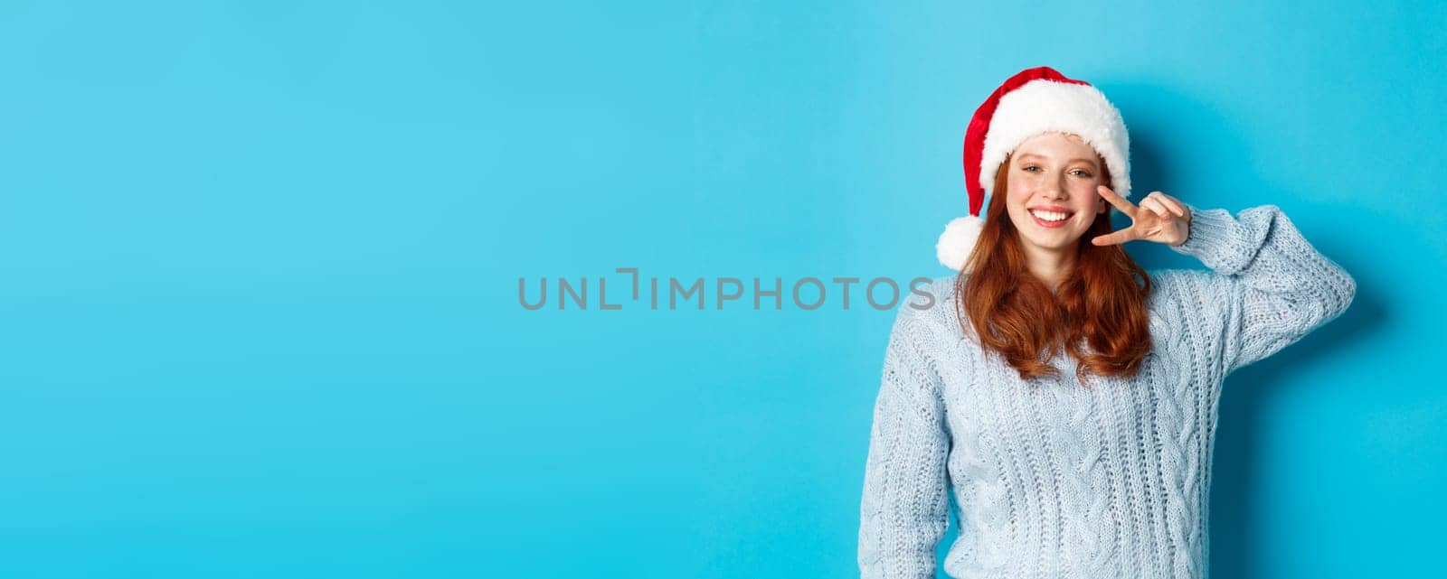 Winter holidays and Christmas Eve concept. Happy teenage girl with red hair, wearing santa hat, enjoying New Year, showing peace sign and smiling, standing over blue background.