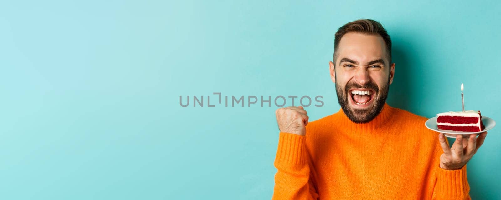 Close-up of happy adult man celebrating birthday, holding bday cake with candle and making wish, standing against turquoise background by Benzoix