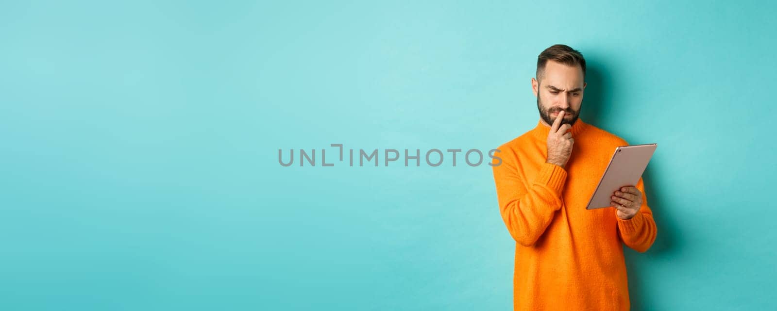 Serious man pondering and looking at digital tablet screen, reading social media, standing over turquoise background by Benzoix