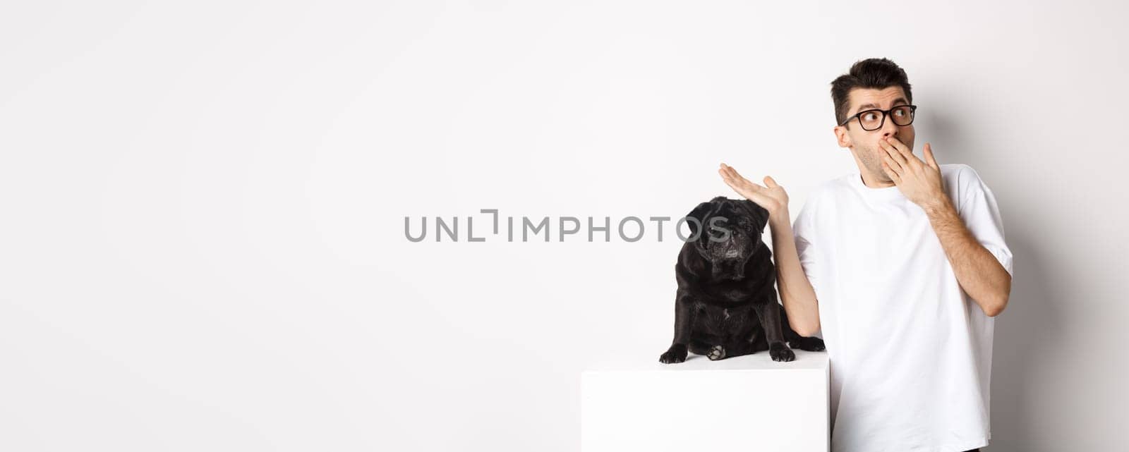 Surprised and shocked man in glasses, standing near cute black dog and staring right at logo, posing over white background.