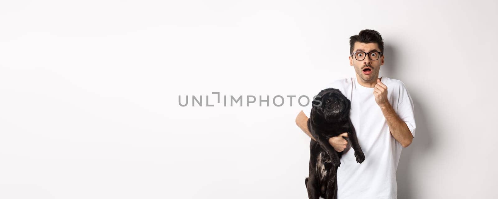 Scared hipster hugging his cute black puppy and staring at camera frightened. Dog owner looking shocked, holding pug, standing over white background.