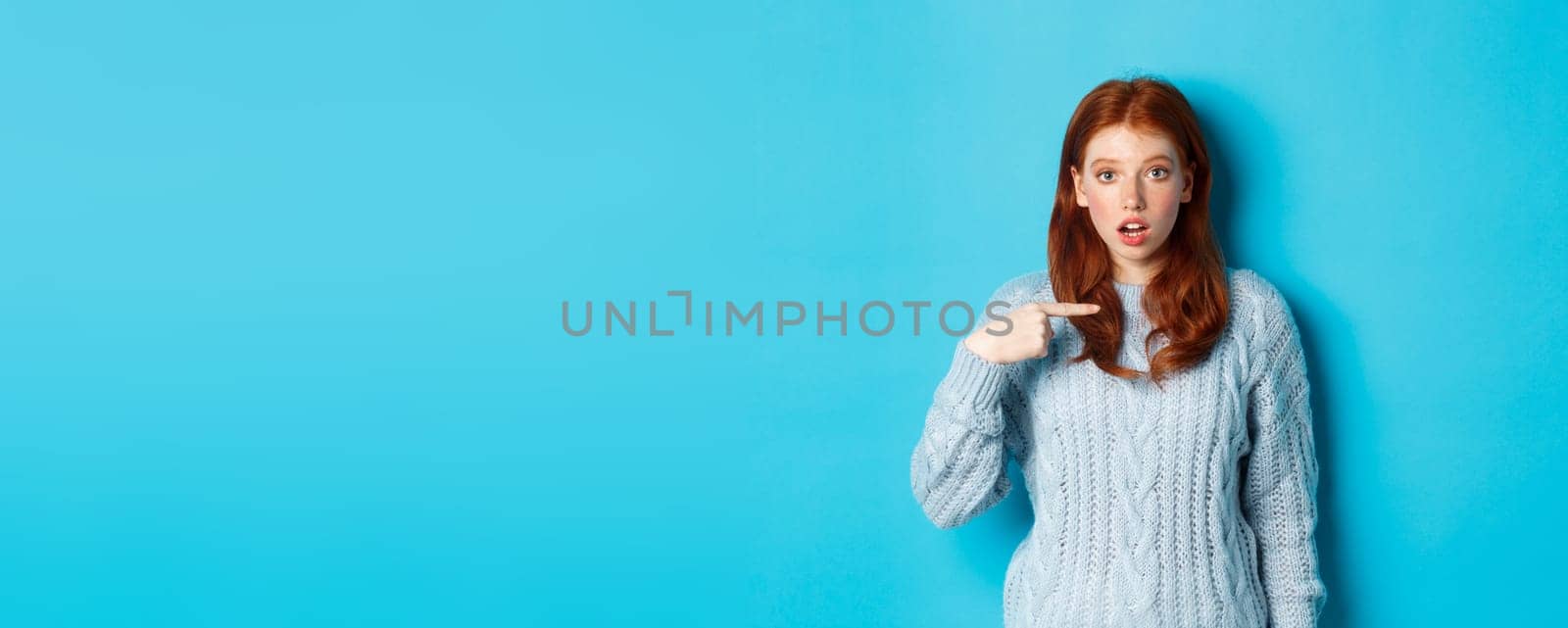 Confused redhead girl pointing at herself, being chosen, standing in sweater against blue background by Benzoix