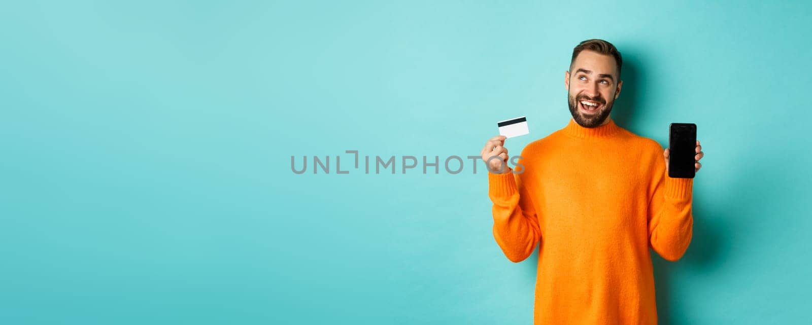 Online shopping. Thoughtful man shop in internet, showing mobile screen and credit card, looking at upper left corner and thinking, standing over light blue background.