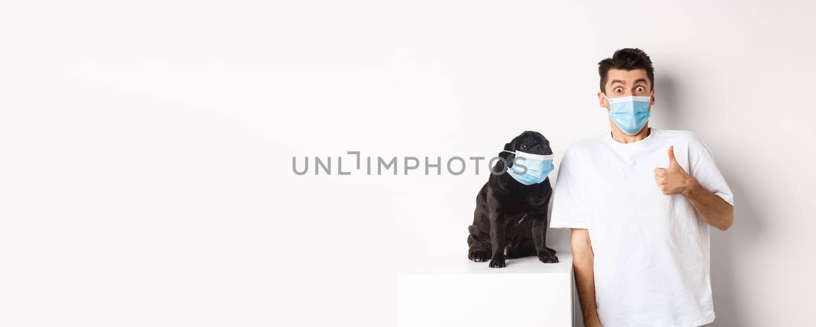 Covid-19, animals and quarantine concept. Image of funny young man and small dog in medical masks, owner showing thumb up in approval or like, white background by Benzoix