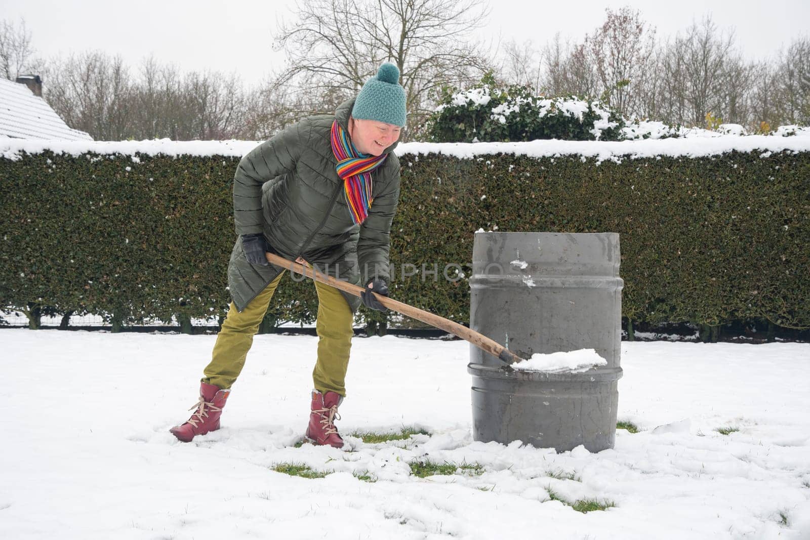 a middle-aged woman is collecting snow in a barrel with a shovel. For further watering plants in a greenhouse, the concept of protecting the environment and conserving natural resources