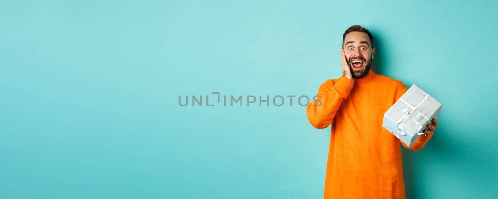 Holidays and celebration concept. Surprised man receiving gift, looking happy at present and smiling, standing over blue background.