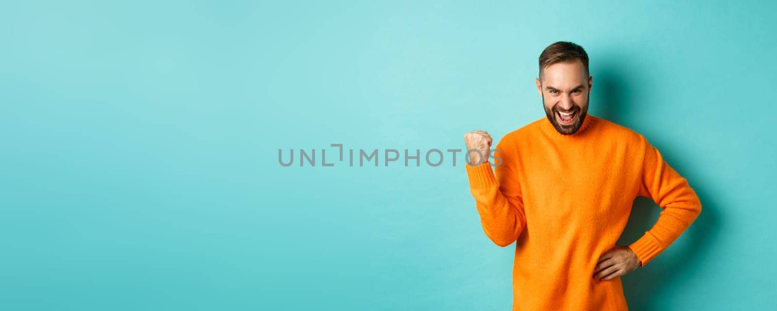 Image of satisfied young man feeling winner, fist pump and saying yes with satisfaction, achieve goal, triumphing while standing over light blue background.