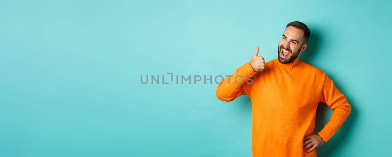 Happy satisfied male model showing thumb up, looking pleased at upper left corner logo, approve and recommend product, standing over light blue background by Benzoix