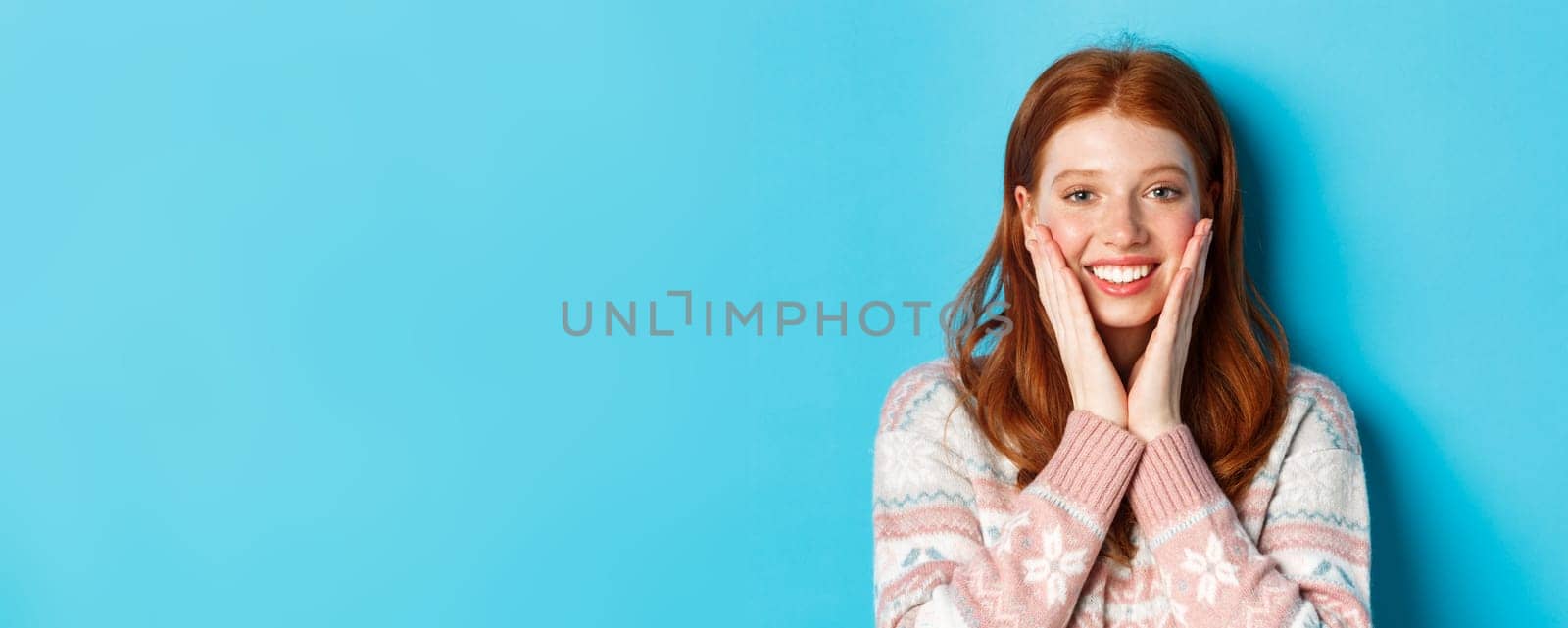 Close-up of cheerful redhead girl blushing, touching cheeks and smiling happy, looking delighted at camera, standing over blue background by Benzoix