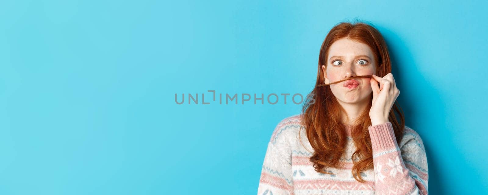 Close-up of silly and funny redhead girl making moustache with hair strand and puckered lips, grimacing against blue background by Benzoix