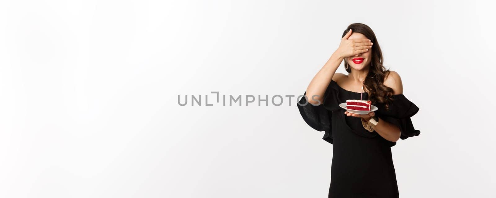 Celebration and party concept. Happy birthday girl in black dress, red lipstick, close eyes and making wish on b-day cake, standing over white background.