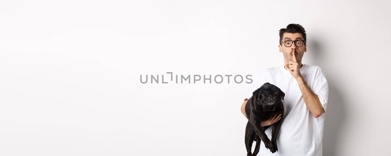 Image of funny young man holding cute black pug dog and shushing at camera, showing hush sign and staring at camera, keeping a secret, standing over white background.