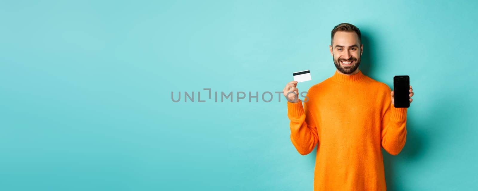 Online shopping. Happy attractive guy showing mobile phone screen and credit card, smiling satisfied, standing over light blue background.