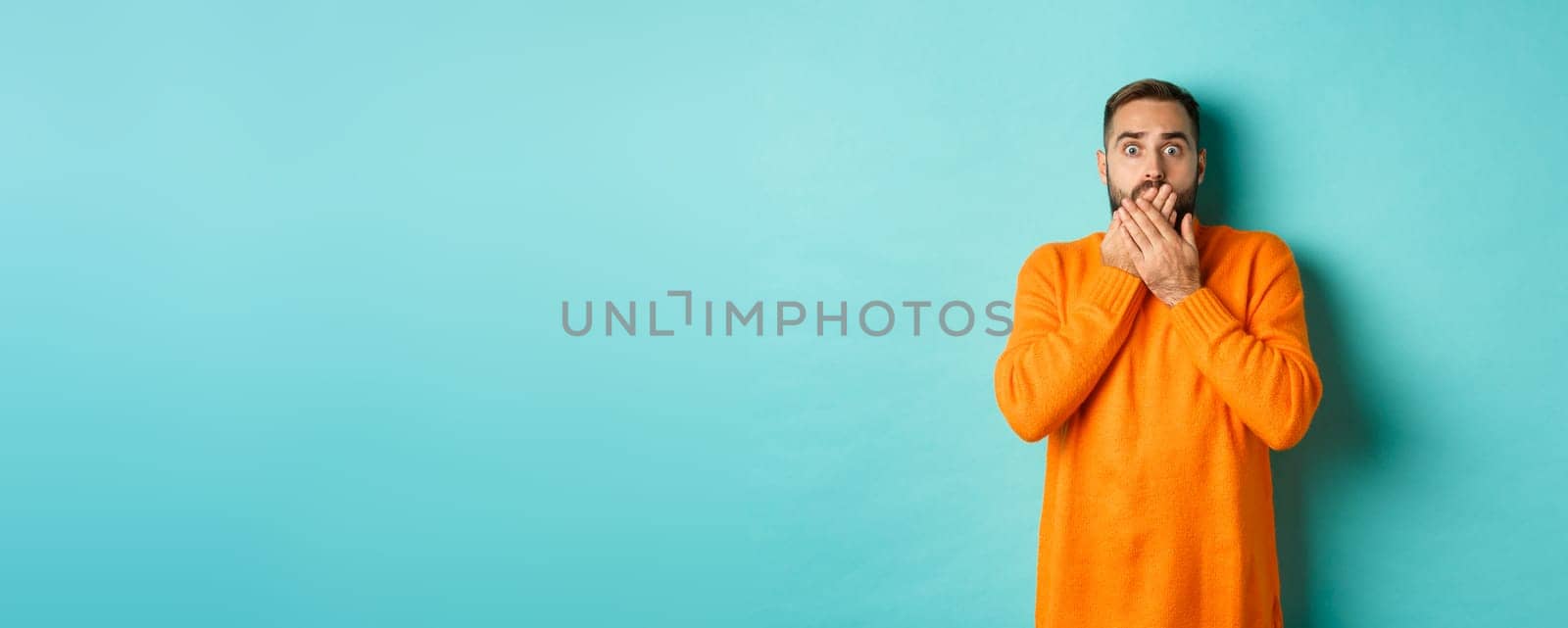 Startled man looking at horrible scene, gasping and stare scared, standing in orange winter sweater, studio background.