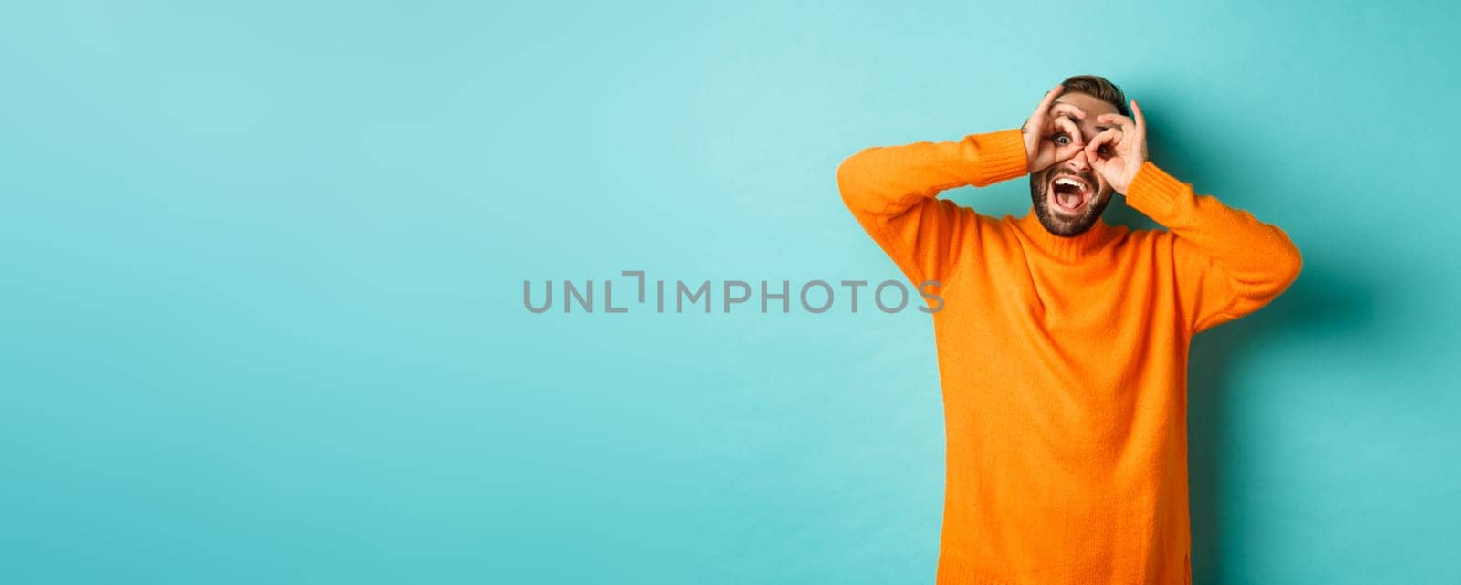 Image of funny and happy guy holding hands on eyes, looking through fake glasses, smiling amused, standing in orange sweater against turquoise background by Benzoix