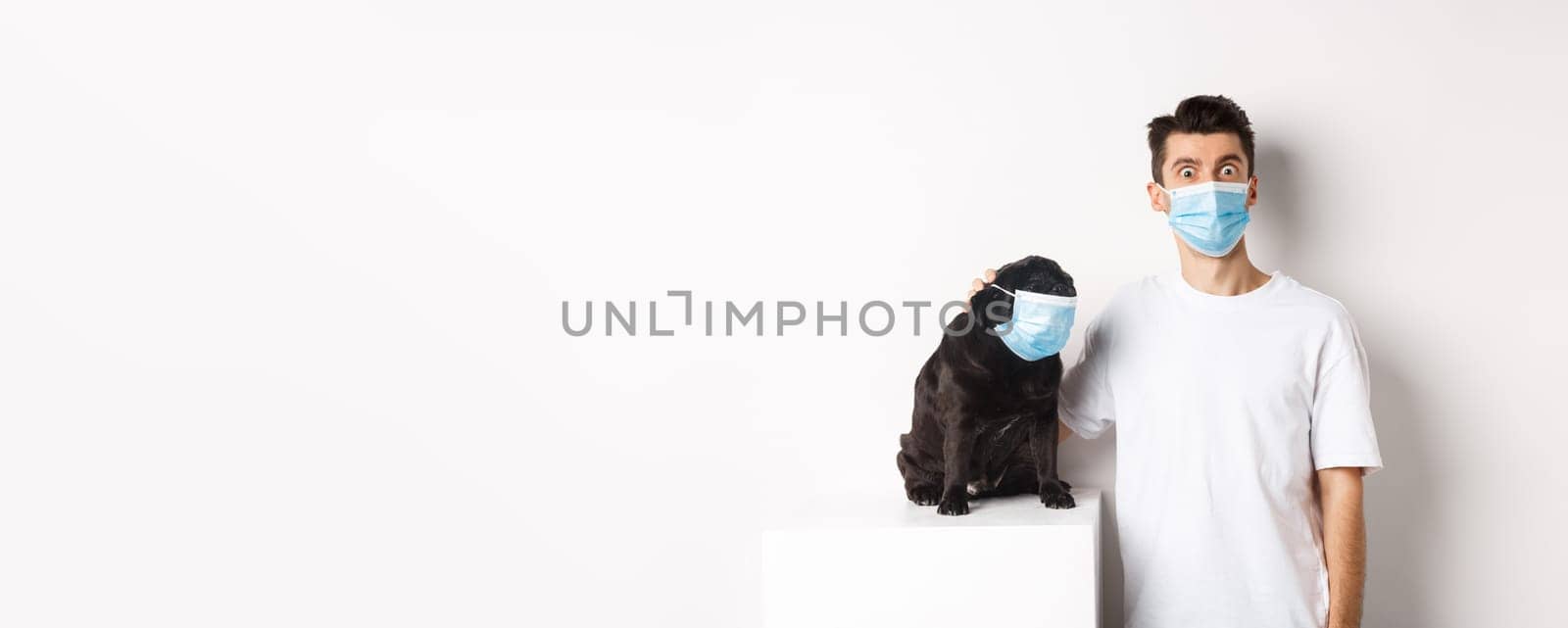 Covid-19, animals and quarantine concept. Image of funny young man and pug in medical masks, staring at camera, standing over white background.