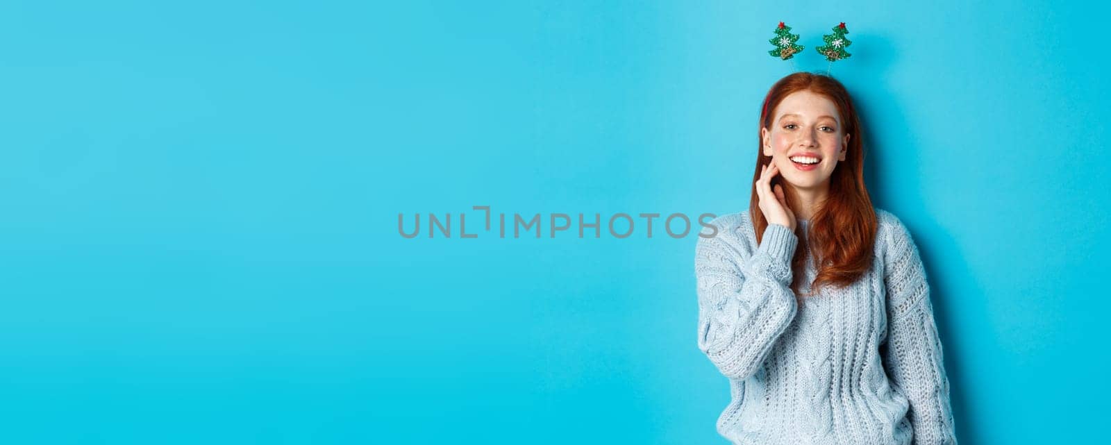 Winter holidays and Christmas sales concept. Beautiful redhead female model celebrating New Year, wearing funny party headband and sweater, smiling at camera.