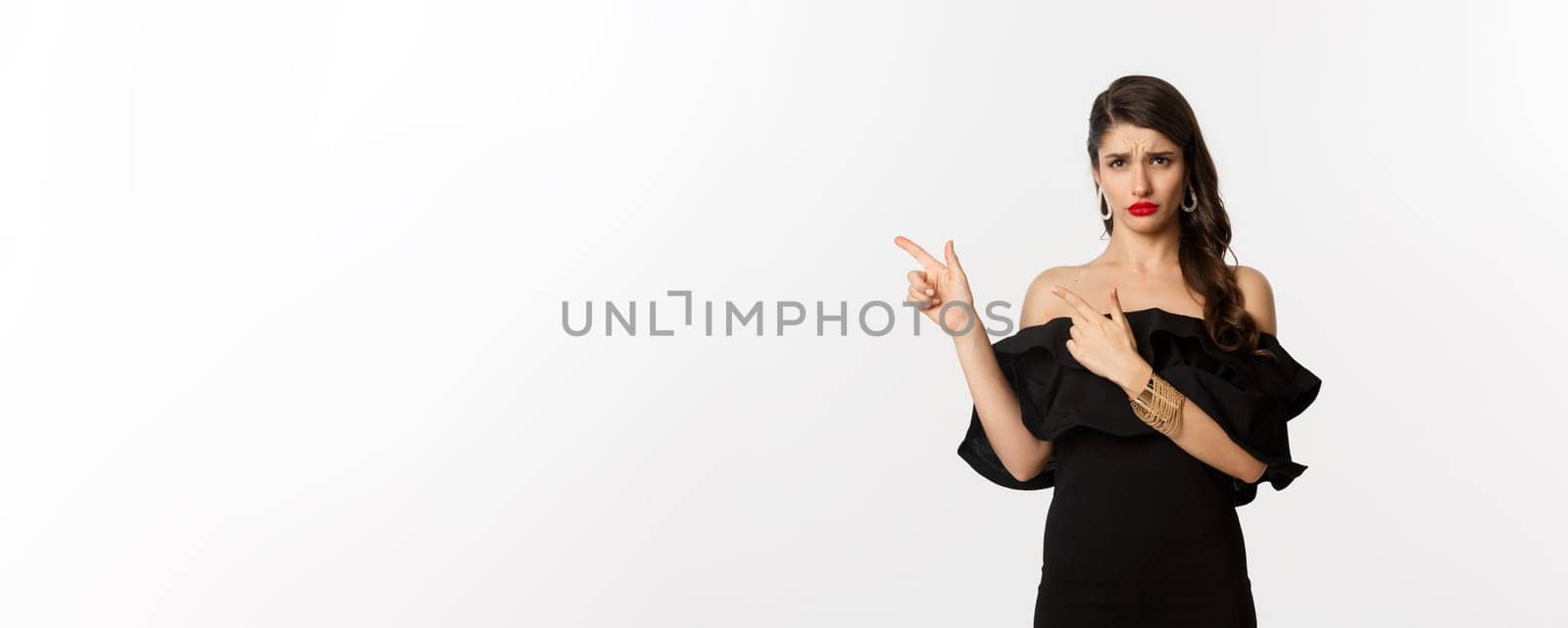 Fashion and beauty. Disappointed glamour woman complaining, wearing black dress, sulking and pointing fingers left at bad offer, white background by Benzoix