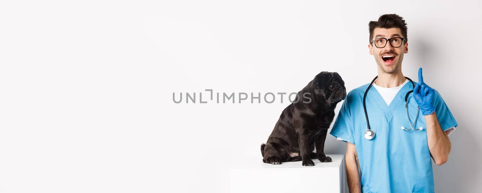 Handsome young doctor at vet clinic pointing finger up and looking amazed, standing near cute black pug dog, white background.